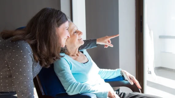 A person in a gray polka-dot sweater leans forward and points out of a window, while another person in a light blue sweater sits and looks in the same direction.